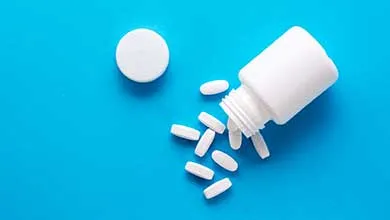 Overhead shot of a white pill bottle, tipped over, spilling white pills onto a bright blue surface, with its cap beside it