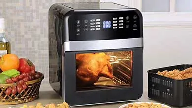 Black digital air fryer oven rotisserie-roasting a whole chicken, surrounded by fried food baskets and fresh fruit on a countertop.