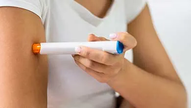 Female in white t-shirt self-administering an injection into their upper arm with a white auto-injector pen.