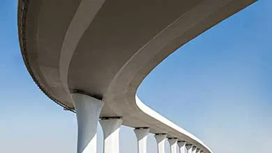 Low-angle shot of a curving concrete overpass, supported by white pillars, against a clear blue sky.