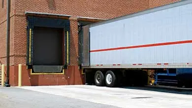 A white semi-truck trailer is parked at a loading dock of a red brick warehouse.