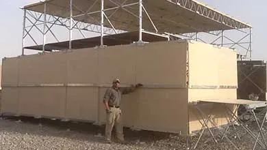 A man stands on gravel beside a large, two-story tan modular building with a metal scaffolding roof.