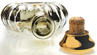 A fluted glass bottle lies on its side beside its stopper, which has a cork base and a shiny gold top with a black pattern.