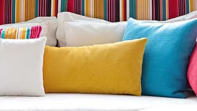 White, yellow, and blue decorative pillows are arranged on a white surface in front of a vibrant, multicolored striped headboard.