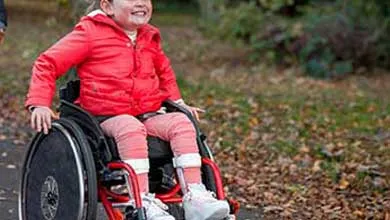 A smiling young girl in a red jacket sits in a wheelchair on an outdoor path covered with fallen leaves.