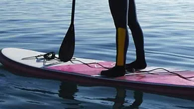 Person standing on a paddleboard on calm open water, holding a paddle with a distant shoreline and clear sky in the background.