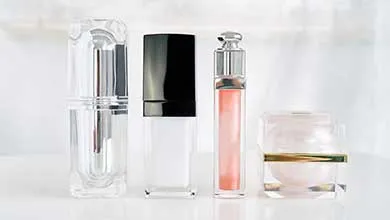 Four cosmetic containers are lined up on a white surface with a bright background. From left to right: a tall clear pump bottle, a rectangular bottle with a black cap, a slim tube with a silver cap containing peach liquid, and a small square jar with a gold band holding white cream.