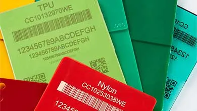 A stack of colorful plastic material ID cards overlapping each other. The front card is red, labeled "Nylon CC10253030WE," featuring a barcode with numbers and a QR code. Behind it, a light green TPU card shows "CC10132970WE" with similar barcode and QR code. Teal, yellow, and cyan cards are partially visible in the background, each displaying alphanumeric product codes, barcodes, and QR codes.