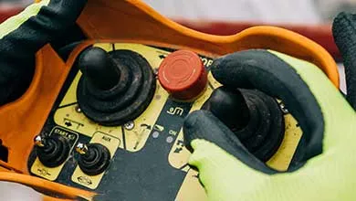 Hands wearing yellow gloves with black fingertips hold an industrial remote control with an orange frame. The panel has two large black joysticks side by side, a red circular button centered between them, and two smaller black knobs labeled "PITCH" in the lower left. The yellow and black surface also includes various switches and markings.