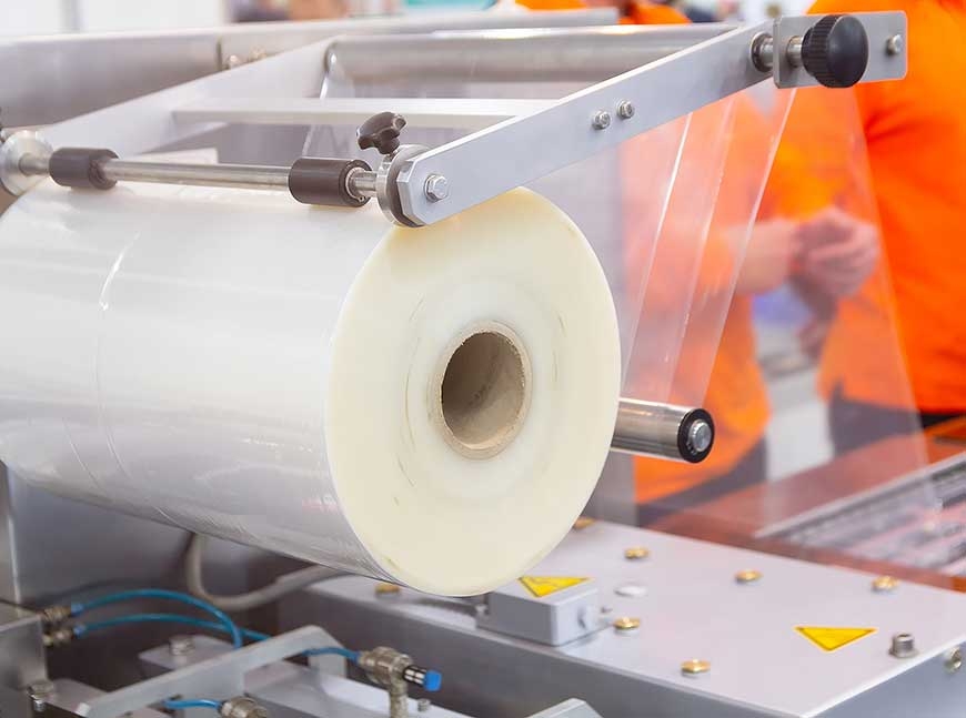 Close-up of a large roll of clear plastic film being fed into an industrial packaging machine.