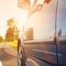 Close-up of a car's side with sunlight flaring over the side mirror and blue sky reflected in the window.